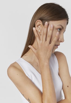 Joven mujer con cabello largo y castaño y una blusa blanca sin mangas cubre la mitad de su rostro con la mano, mostrando un pequeño pendiente de perno y un tatuaje.
