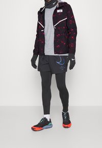 Zwarte jas met rood en roze patroon, grijs shirt, zwarte handschoenen, zwarte shorts met blauw logo, zwarte en oranje hardloopschoenen.