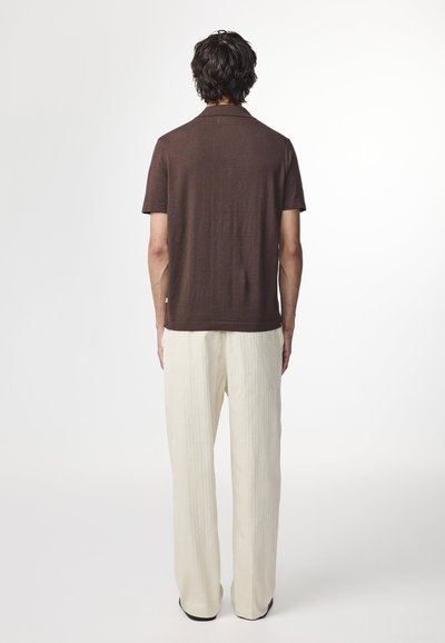 Polo shirt marron à manches courtes associé à un pantalon large crème, présentant des lignes texturées verticales et une coupe décontractée.