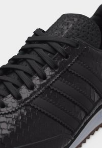 Gros plan sur une basket noire texturée avec des lacets noirs, présentant trois bandes mates sur le côté et un bord de semelle noir rainuré.