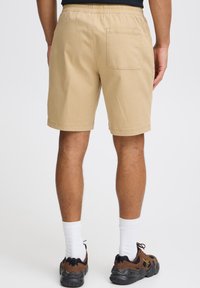 Kaki värvi lühikesed püksid elastse vöökohaga ja tagataskuga. Kangas näib sile ning klassikalise lõikega, sobitatuna pruunide ja mustade tossudega.