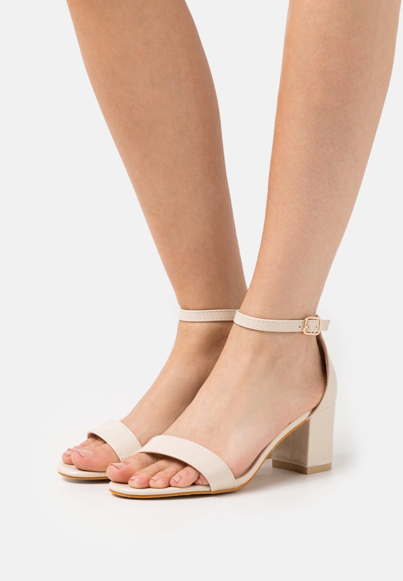 Sandales beige à bride cheville avec une large bande à l'avant, une finition brillante et un talon épais. Les brides sont ornées d'une boucle carrée.