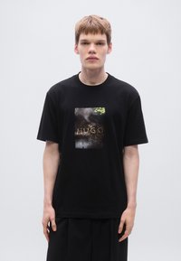 Μαύρο βαμβακερό T-shirt με αντανακλαστικό ορθογώνιο  γράφημα που φέρει τη λέξη "HUGO." Κοντά μανίκια, άνετη γραμμή, μινιμαλιστικός σχεδιασμός.