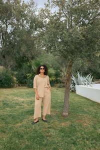 Frau mit lockigem Haar, beige Outfit und schwarzen Schuhen steht auf Gras neben einem Baum in einem Garten mit dichtem Laub.