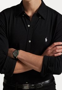 Svart langermet polo skjorte med teksturert stoff, med en hvit brodert logo, og et svart klokke med rund urskive og lærrem.