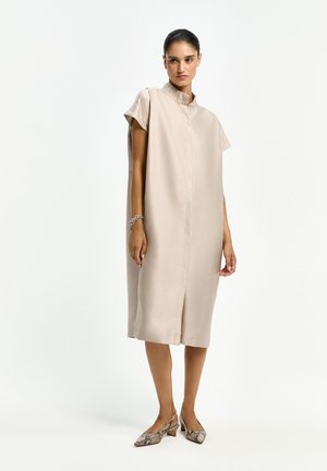 Frau steht und trägt ein locker sitzendes beiges Midikleid mit Stehkragen, kurzen Ärmeln, Schlangenmuster-Schuhe mit niedrigem Absatz und ein silbernes Armband.