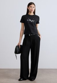 Μαύρο βαμβακερό T-shirt με λευκό γραφικό κείμενο, συνδυασμένο με μαύρο παντελόνι ψηλόμεσο, φαρδύ και μικρή μαύρη τσάντα με κορδόνι.