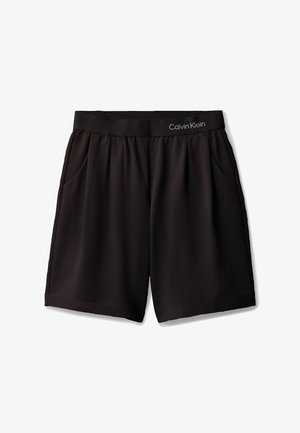 Junges Mädchen steht mit den Händen in den Taschen, trägt ein schwarzes ärmelloses Oberteil, schwarze Shorts und schwarze Loafers vor grauem Hintergrund.