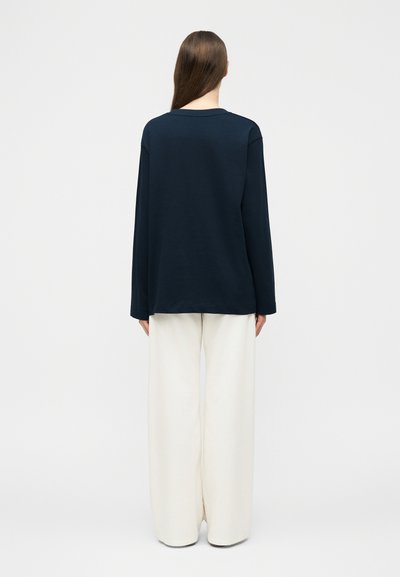 Γυναίκα που στέκεται με την πλάτη γυρισμένη, φοράει μπλε τοπ μακρυμάνικο και άσπρο παντελόνι φαρδύ, μπροστά σε απλό ανοιχτόχρωμο φόντο.