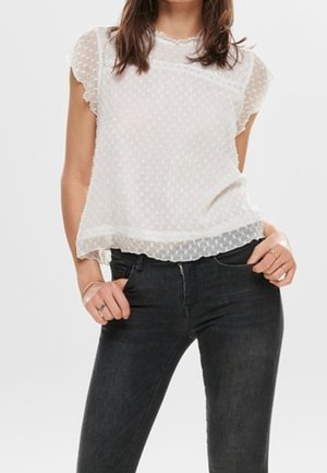 Vrouw met een doorschijnende witte blouse met korte mouwen en polka dots, ingestopt in zwarte nauwsluitende jeans, handen rustend bij de taille.