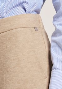 Beige Stoffhose mit glatter Textur, ausgestattet mit einer Seitentasche und einem minimalistischen silbernen Akzentknopf, kombiniert mit einem blau gestreiften Hemd.