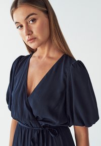 Navy blue wrap dress with a deep V-neck, pleated texture, and puff sleeves. Tied at the waist with a matching belt for adjustable fit.