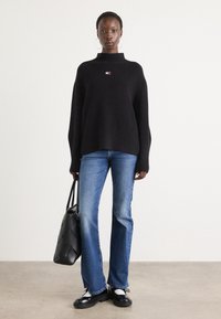 Svart ribbestrikket genser med høy krage og liten brodert logo, sammen med blå utsvingte jeans og svart totebag. Svarte og hvite sko.