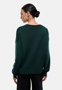 Pull en maille vert foncé avec une coupe décontractée, un col et des poignets côtelés. Présente des épaules tombantes et une longueur légèrement courte.