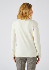 Maglione crema chiaro con maniche lunghe, orlo a coste e texture liscia. Indossato sopra pantaloni grigi a scacchi, mostrato da dietro.