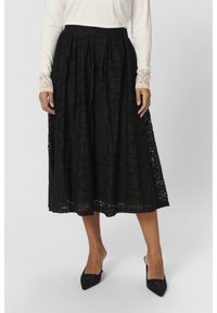 Zwarte midi rok met een intrigerend kantontwerp, geplooide stof en een gegolfde zoom. Gestyleerd met een crèmekleurige top en puntige zwarte hakken.