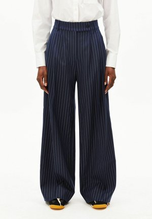 Personne portant un pantalon large bleu marine à fines rayures, une chemise blanche à manches longues et des chaussures noires avec des semelles orange.