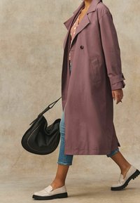 Femme marchant, portant un trench mauve, un jean bleu raccourci, des mocassins beiges, portant un sac à main noir sur un fond neutre.