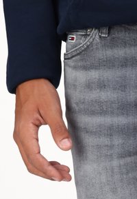 Levá ruka osoby blízko kapsy šedých džín s malým logem Tommy Hilfiger, oblečená v tmavě modrém tričku s dlouhým rukávem.