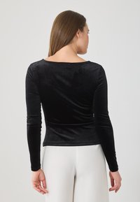 Žena s dugom smešnom kosom, obučena u uski crni duksericu dugih rukava i bele pantalone, okrenuta leđima prema jednostavnoj svetloj pozadini.