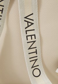 Beige läderväska med en texturerad yta, med ett framträdande vitt band med svart "VALENTINO"-text.