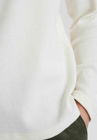 Weißer, strukturierter Strickpullover mit Rundhalsausschnitt und langen Ärmeln, der ein geripptes Muster aufweist. Der Saum ist leicht nach oben gerollt.