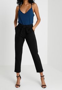 Donna che indossa un top blu con bordo a scallop, pantaloni neri a vita alta tipo paperbag con cintura e tacchi ankle-strap neri, in piedi con le mani nelle tasche.