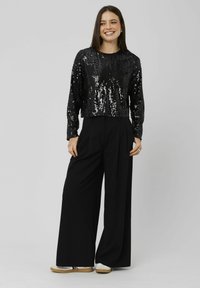 Top en sequins noir court avec des manches longues, associé à un pantalon large noir taille haute. Le mannequin se tient avec les mains sur les hanches.