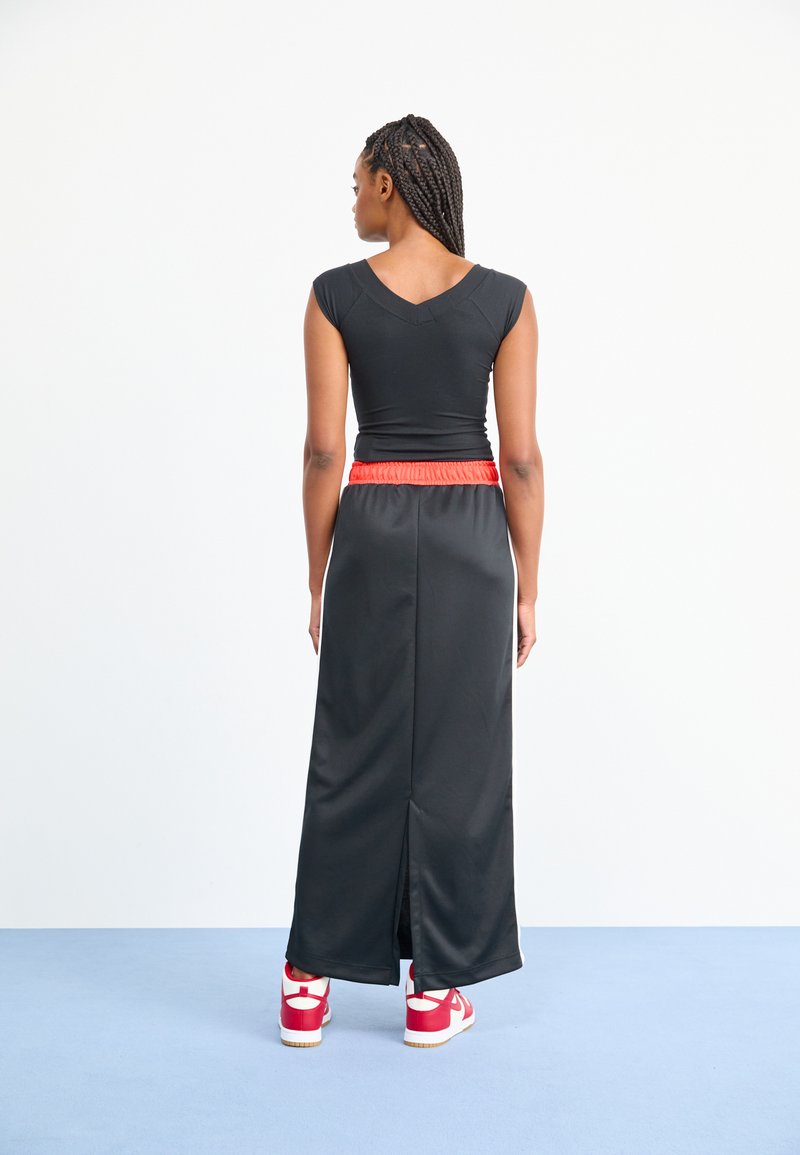 Top atlético negro con escote en V, combinado con una falda larga negra que presenta una cinturilla elástica roja. La modelo lleva zapatillas rojas.