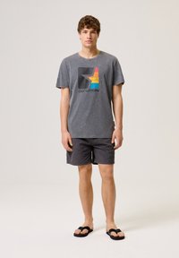 Szary, krótki t-shirt z kolorową grafiką i logo, w zestawieniu z ciemnymi szortami oraz czarnymi klapkami na neutralnym tle.