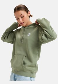 Sudadera verde hecha de material suave, con un bolsillo frontal y capucha con cordón. Presenta un pequeño logo de sol blanco en el pecho. Corte relajado.