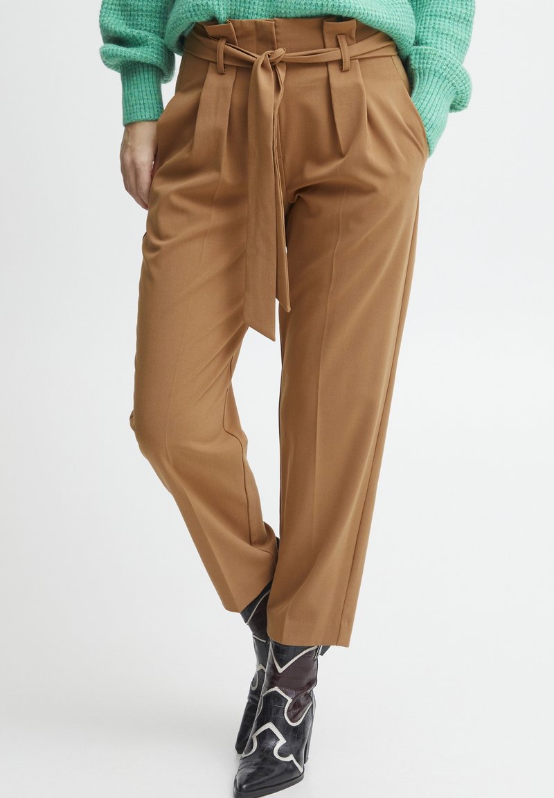 Pantalon marron sur mesure à taille haute, avec des plis et une ceinture en tissu assorti. Porté avec des bottes noires et marron à motif.