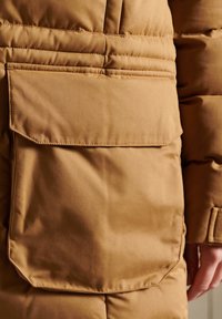 Le tissu de la veste isolante marron présente une grande poche avant avec un rabat, une texture lisse et des coutures matelassées horizontales.