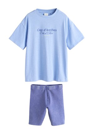 Jasnoniebieski T-shirt z krótkim rękawem z napisem "Cap d’Antibes Côte d’Azur" zestawiony z fioletowymi spodenkami do kolan.