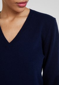 Mörkblå V-ringad tröja tillverkad av mjukt stickat material; har långa ärmar och en figurnära design. Den texturerade tyget ger dimension.