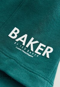 Detailný záber na tyrkysovú textíliu s bielym potlačeným textom: "BAKER BY TED BAKER Londýn, Spojené kráľovstvo," zobrazuje detaily šitia.