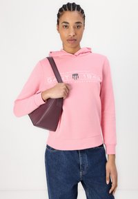 Rosa bomullshoodie med kengurulomme og hvit logo tekst, kombinert med mørkeblå jeans og en liten bordeaux skulderveske.
