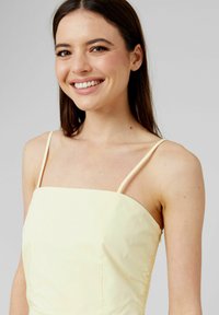 Robe jaune pâle avec fines bretelles, décolleté carré et tissu texturé. Présente des fronces sur les côtés et une silhouette cintrée.