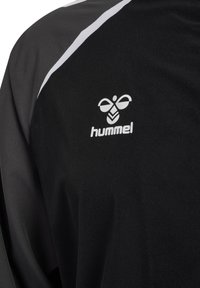 Primer plano de chaqueta deportiva negra y gris con logo blanco de Hummel y una franja blanca diagonal en el hombro.