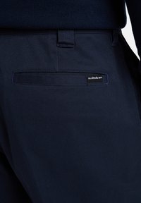 Námornícke modré nohavice z odolného materiálu so štíhlym strihom, s jednou zadnou vreckom a štítkom Quiksilver na okraji vrecka.