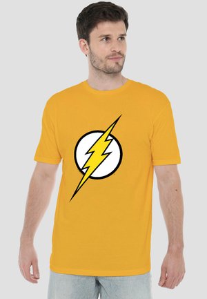 Mann trägt gelbes T-Shirt mit großem weißem Kreis mit schwarzem Rand und gelbem Blitzsymbol, schaut nach rechts, grauer Hintergrund.