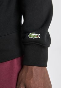 Black sweatshirt featuring a ribbed cuff and a green crocodile logo embroidered on the sleeve. The fabric appears soft and textured.