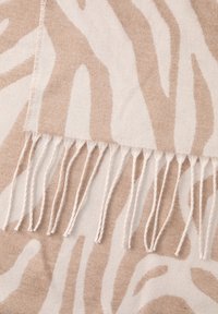 Beige tæppe med zebramønster og frynser i kanten, lavet af blødt vævet materiale med en diskret tekstur og kontrasterende cremefarvede striber.