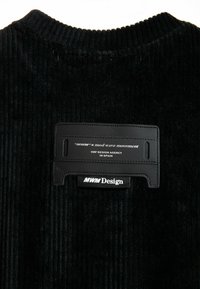 Tela de pana negra con una textura acanalada. Presenta una etiqueta de cuero negro con texto y detalles del logotipo en blanco, ubicada en el cuello.