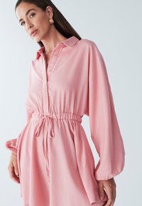 Rozā krekla kleita ar apkakli, elastīgu jostasvietu un garām pūstām piedurknēm. Mīksta auduma ar pogu detaļām un auklas dekoru jostasvietā.