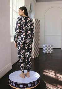 Femme aux longs cheveux noirs se tient pieds nus sur une plateforme ronde, portant un ensemble coordonné en imprimé floral sombre dans une pièce lumineuse avec des piliers à damier.