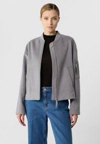 Frau in einer hellgrauen Bomberjacke mit Reißverschluss, schwarzem Oberteil und blauen Jeans, die vor einem einfarbigen hellen Hintergrund steht.