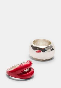 Rød emaljeret ring formet som læber ved siden af en tekstureret, rund sølvring med en blank finish.
