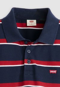 Ναυτικό πόλο μπλουζάκι με κόκκινες και λευκές οριζόντιες ρίγες. Διαθέτει γιακά, τρεις κουμπιά μπροστά και ετικέτα με το λογότυπο της Levi's.