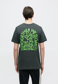 T-shirt de algodão cinzento escuro com uma grande imagem verde nas costas, com o texto "JUNTOS NAVEGAMOS" e um desenho de borboleta. Mangas curtas.